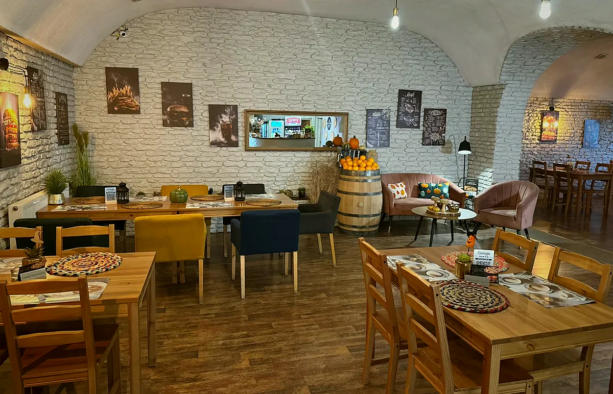 Druga sala z drewnianymi stołami, kolorowymi fotelami, beczką z pomarańczami i kącikiem z sofą